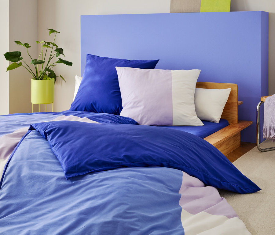 Ein Bett mit blau-lila Bettwäsche und Kissen steht vor einer blauen Wand, daneben eine Pflanze und ein Stuhl mit einer Decke.