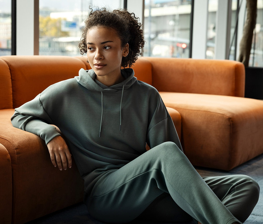 Frau sitzt auf dem Boden, angelehnt an ein orangefarbenes Sofa, und trägt einen grünen Hoodie und eine Jogginghose.
