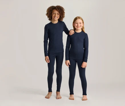 Zwei lächelnde Kinder, ein Junge mit lockigem braunem Haar und ein Mädchen mit glattem blondem Haar, stehen nebeneinander in passenden marineblauen Langarmshirts und Leggings. Der größere Junge hat seinen Arm auf der Schulter des Mädchens; beide sind barfuß.