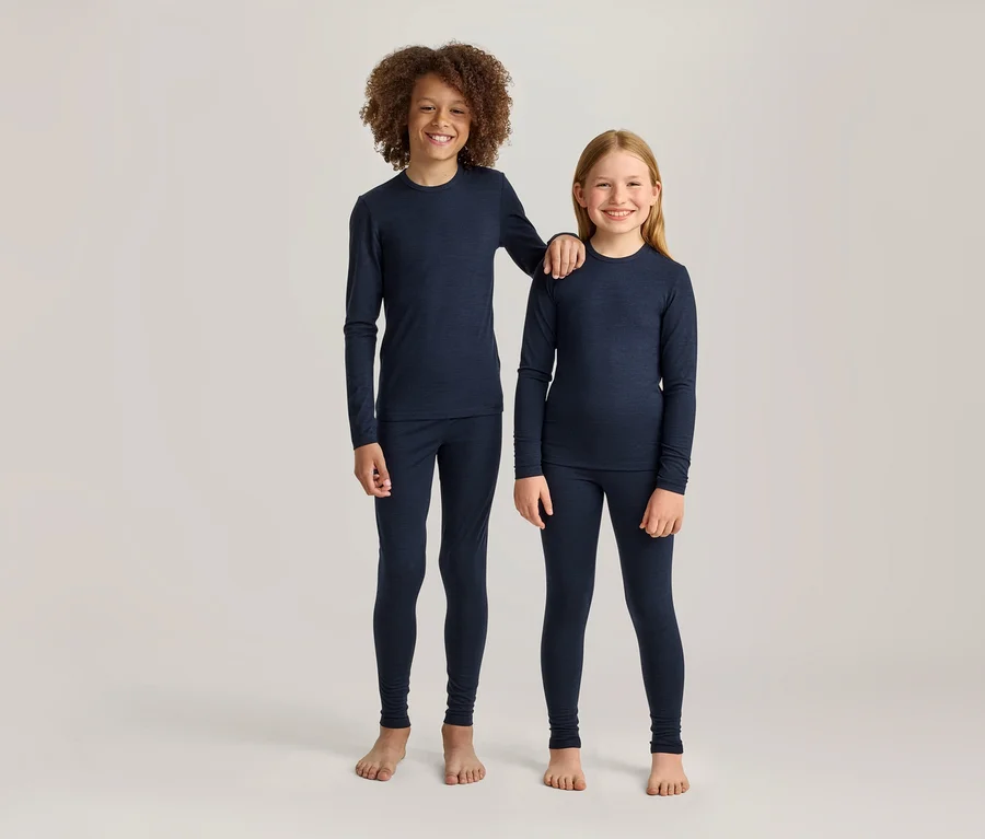 Zwei lächelnde Kinder, ein Junge mit lockigem braunem Haar und ein Mädchen mit glattem blondem Haar, stehen nebeneinander in passenden marineblauen Langarmshirts und Leggings. Der größere Junge hat seinen Arm auf der Schulter des Mädchens; beide sind barfuß.