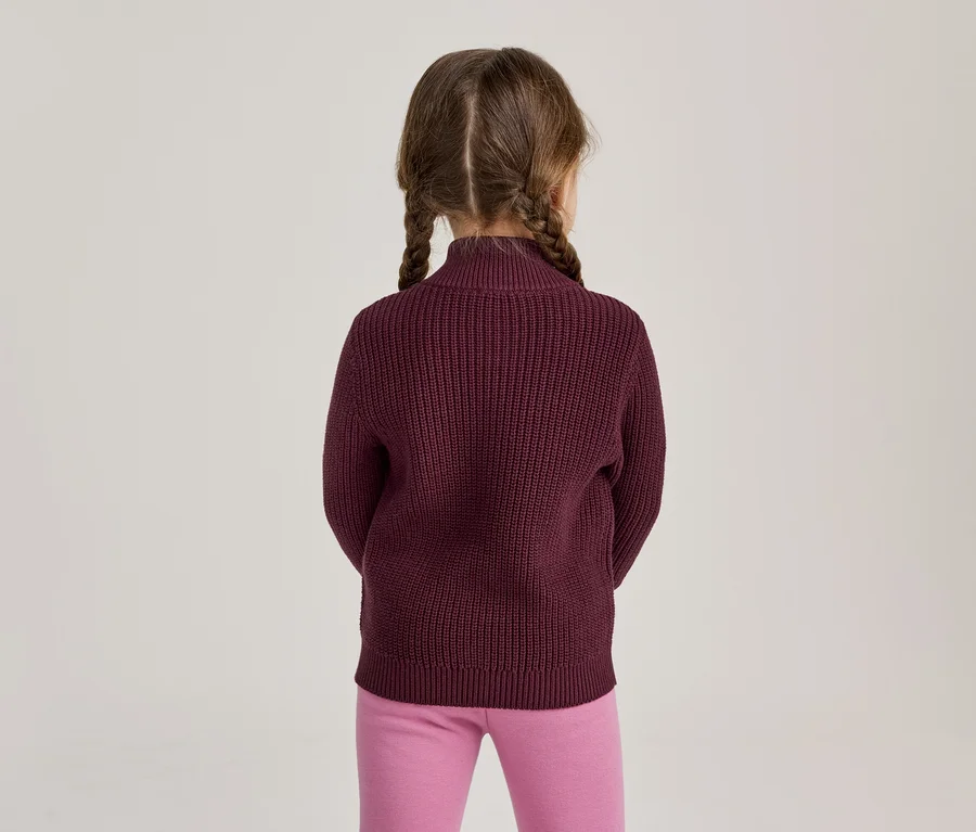 Mädchen von hinten gesehen, trägt einen burgunderroten Strickpullover und pinke Leggings, die Haare sind zu zwei Zöpfen geflochten.