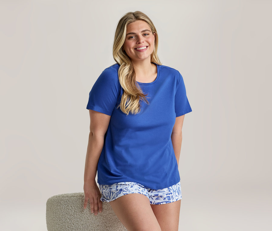 Lächelnde Frau, die ein royalblaues Shorty-Pyjama-Set mit unifarbenem Oberteil und gemusterten Shorts trägt und sich an einen Hocker lehnt.