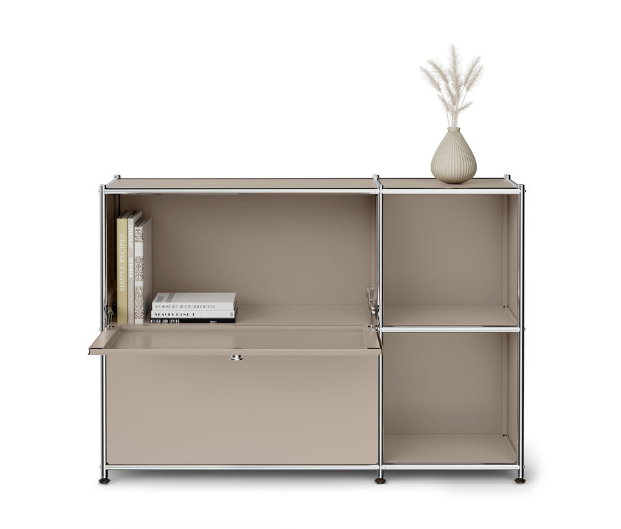 Ein beiges, modulares Aufbewahrungsmöbel mit Chromrahmen. Die linke Seite hat ein offenes Fach mit mehreren Büchern und eine geöffnete Klapptür, die eine Schreibtischfläche bildet. Die rechte Seite weist zwei offene Fächer auf. Eine kleine, gerippte beigefarbene Vase mit getrockneten Gräsern steht oben auf dem Möbel.