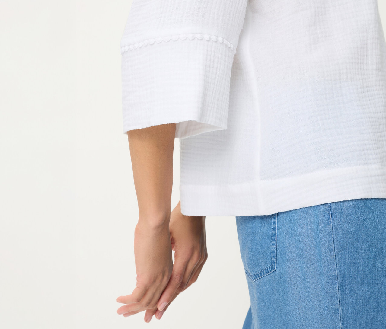 Nahaufnahme einer Person in einer weißen Olsen Webbluse mit strukturierter Oberfläche und dekorativer Borte an den Ärmeln, kombiniert mit hellblauen Jeans. Die Hände der Person sind gefaltet.