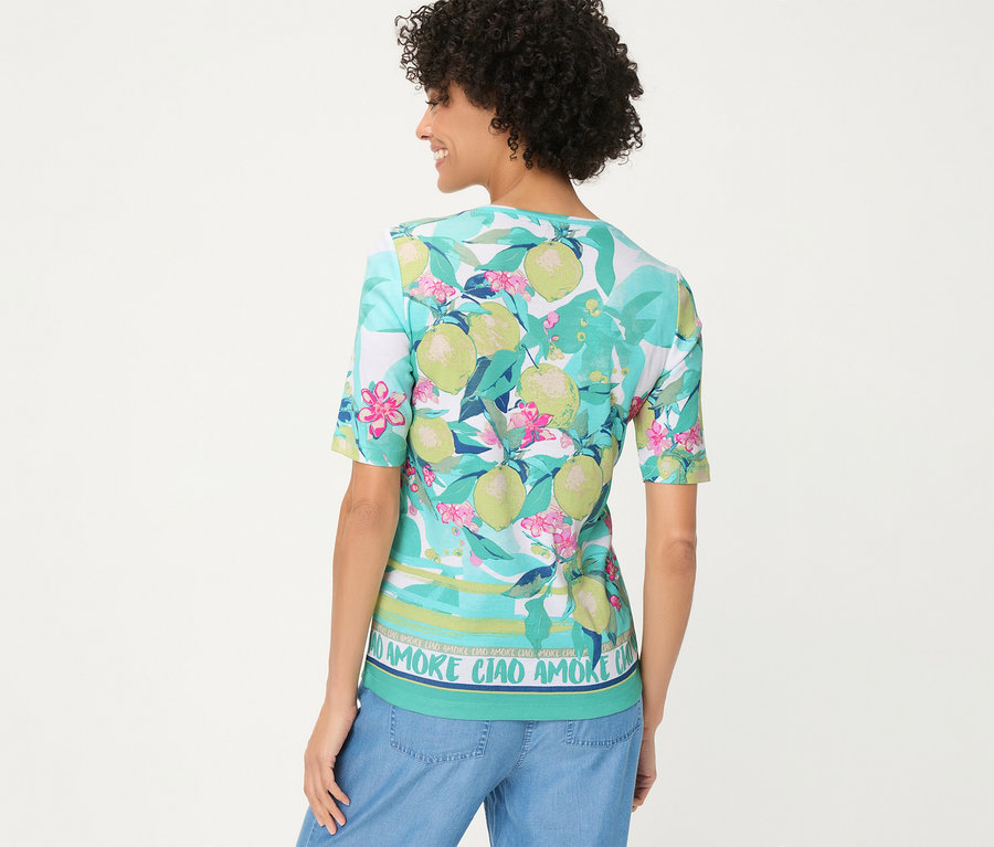 Frau von hinten in einem Olsen T-Shirt mit Zitronen-, Blätter- und pinkem Blumenmuster, gestreiftem Saum mit „AMORE CIAO“-Schriftzug und blauen Jeans.