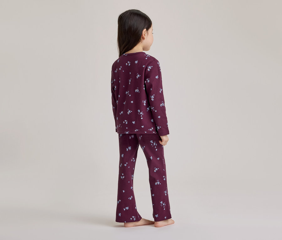 Junges Mädchen von hinten gesehen, trägt einen dunkelvioletten gerippten Pyjama mit hellem Blumenmuster, barfuß stehend.