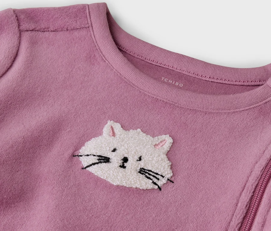 Rosa Fleece-Babyschlafanzug mit einer weißen, flauschigen Katzenstickerei mit schwarzen Schnurrhaaren und rosa Innenohren. Ein diagonaler Reißverschluss und Schulterdruckknöpfe sind sichtbar, 'TCHIBO' ist am Kragen aufgedruckt.
