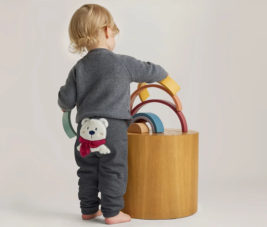 Kleinkind von hinten, in grauem Pullover und Hose mit Eisbär-Applikation, spielt mit bunten Holz-Regenbogen-Stapelspielzeugen auf einem Holzhocker.