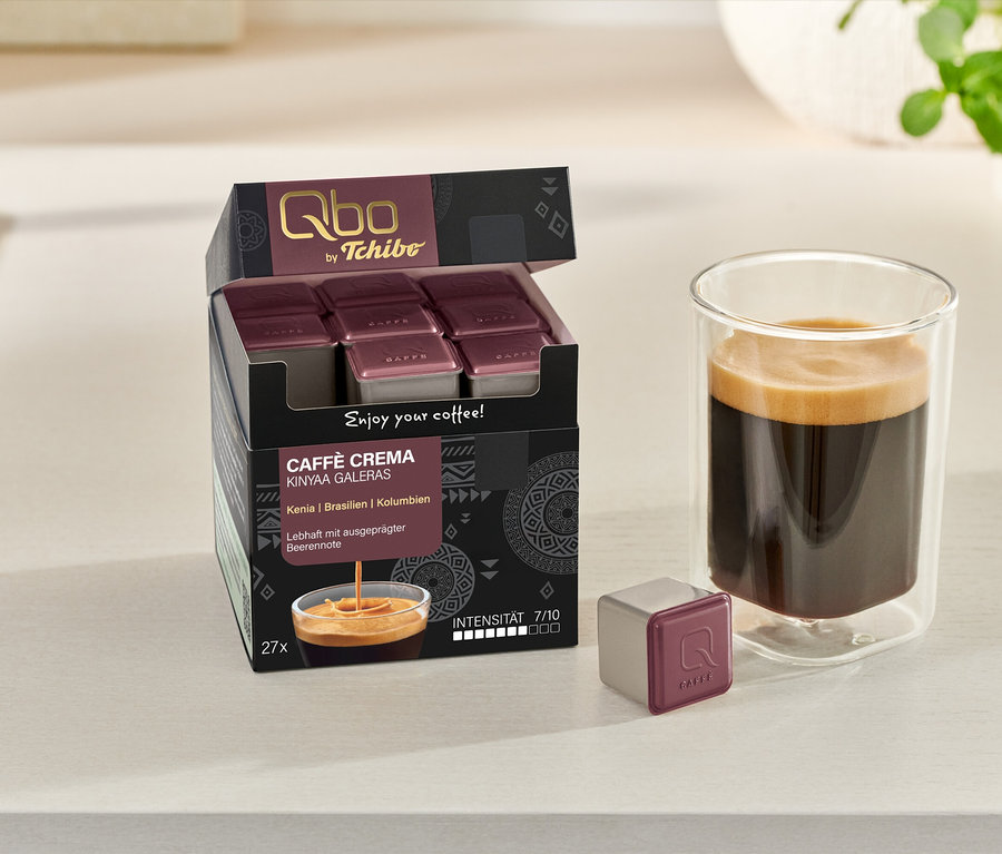 Geöffneter Karton Qbo Caffè KINYAA GALERAS mit Kaffeetasse und Kapsel auf Tisch.