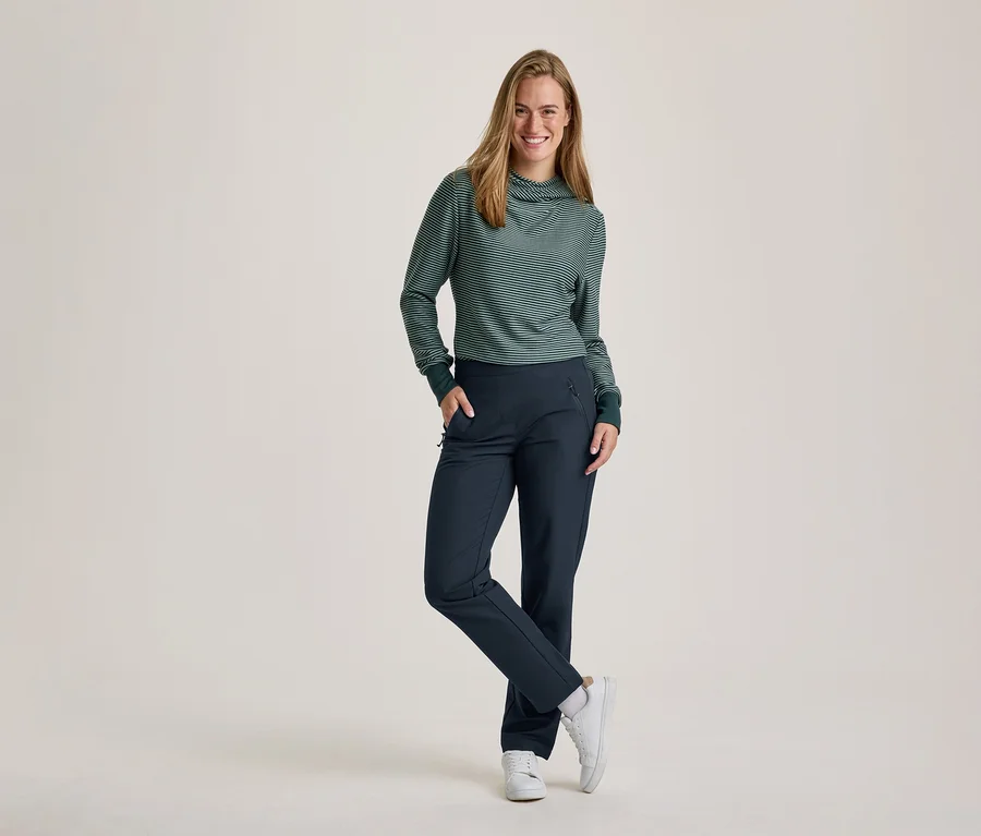 Eine lächelnde Frau mit langen Haaren steht mit einer Hand in der Tasche und einem gekreuzten Bein. Sie trägt ein grün-weiß gestreiftes Langarmshirt mit Rollkragen und dunkelblaue Hosen sowie weiße Turnschuhe.