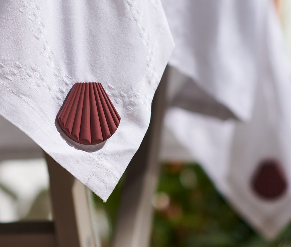 Tischkante mit weißer Tischdecke, beschwert mit braunem 4 Tischdeckenbeschwerer in Muschelform.