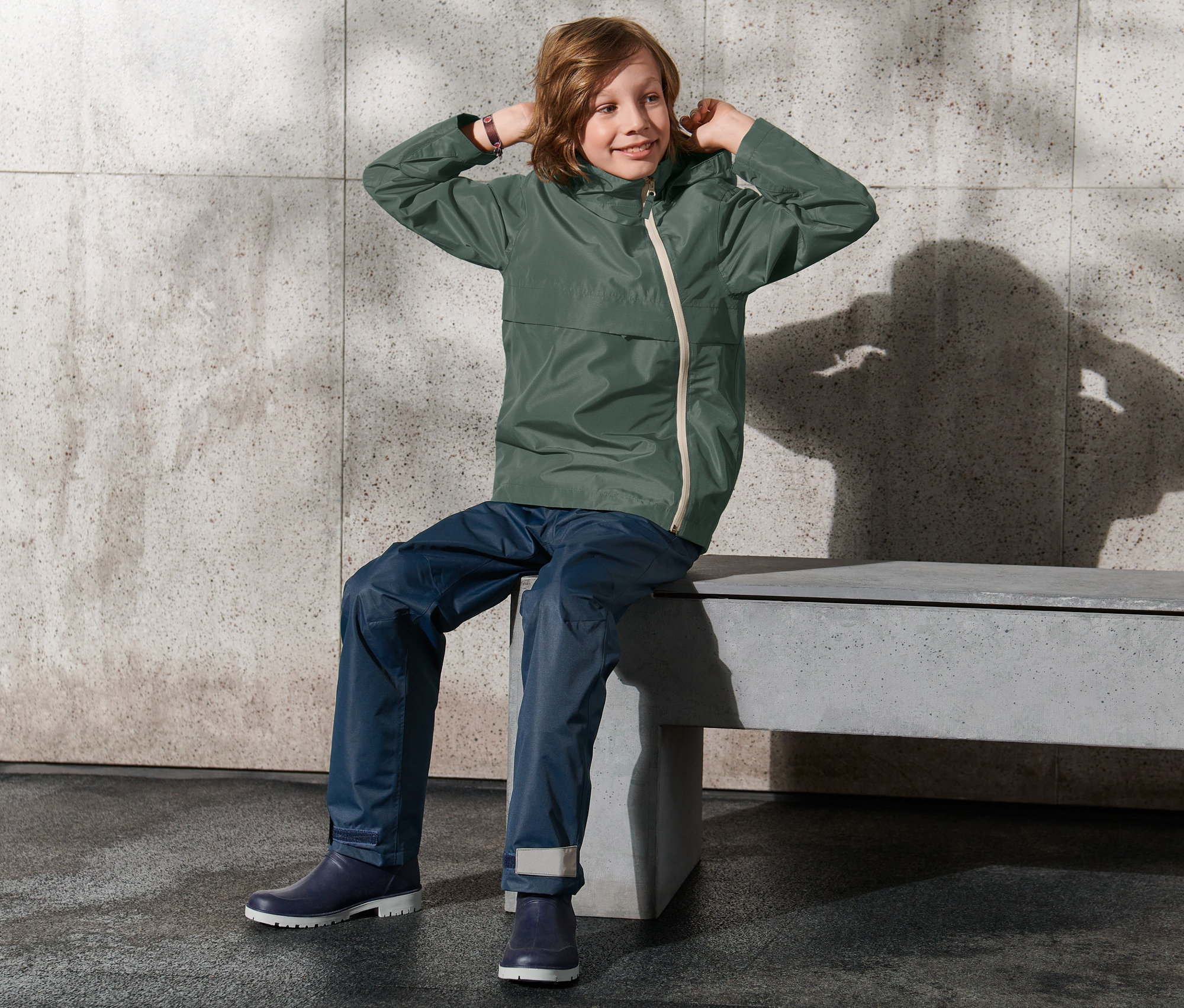 Junge sitzt auf Bank und zieht die Kapuze seiner Allwetterjacke hoch.