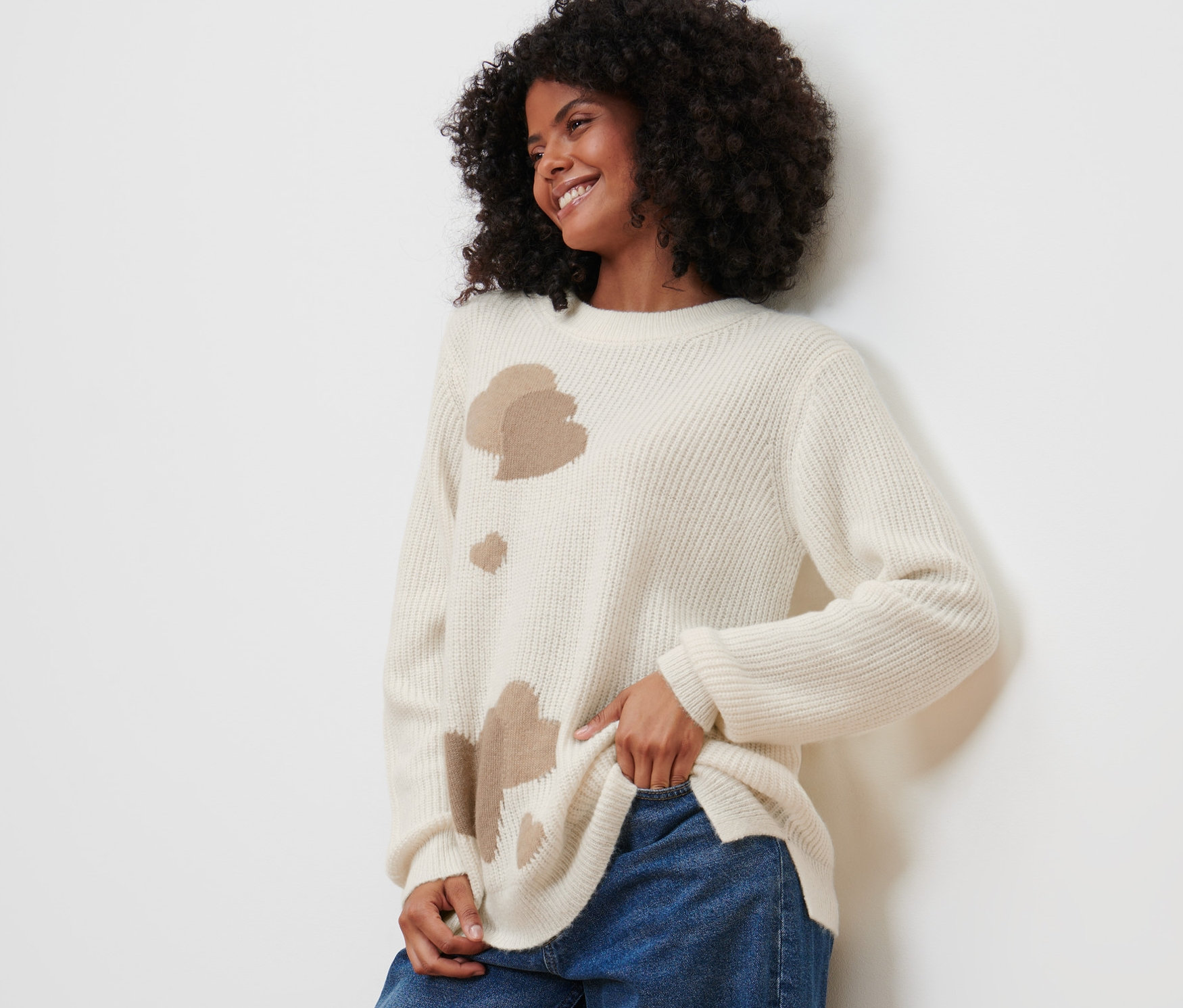 Frau mit lockigem Haar posiert in einem Strickpullover mit Herz-Jacquard und blauen Jeans.