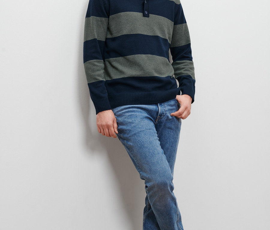 Mann an eine Wand gelehnt, trägt einen gestreiften Pullover und Jeans.