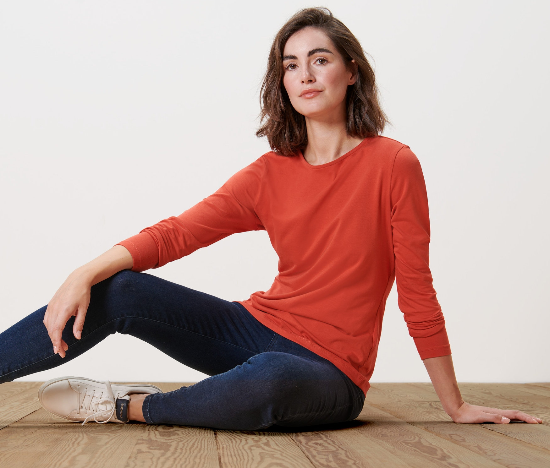 Frau sitzt auf Holzboden und trägt ein orangefarbenes Langarmshirt.