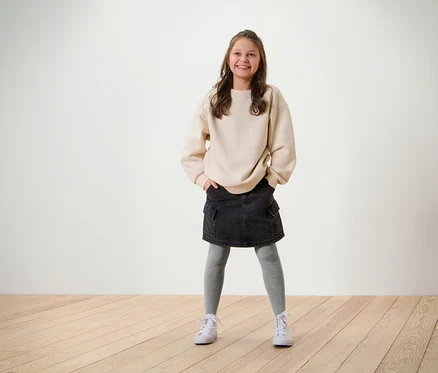 Lächelndes Mädchen posiert in beigem Sweatshirt, schwarzem Rock und grauer 5 Paar Kinder-Strumpfhosen.