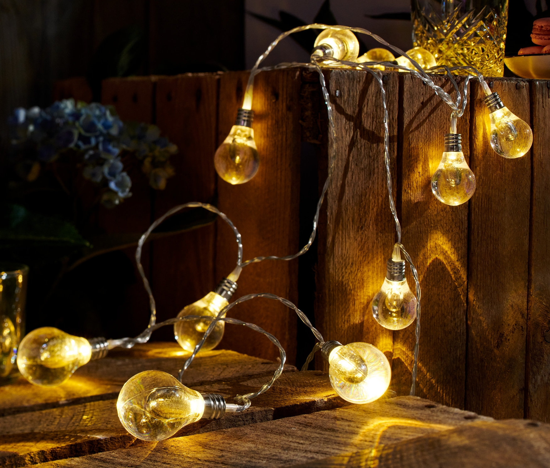 Eine LED-Lichterkette liegt auf einem Holztisch, im Hintergrund ein Glas mit Getränk und Blumen.