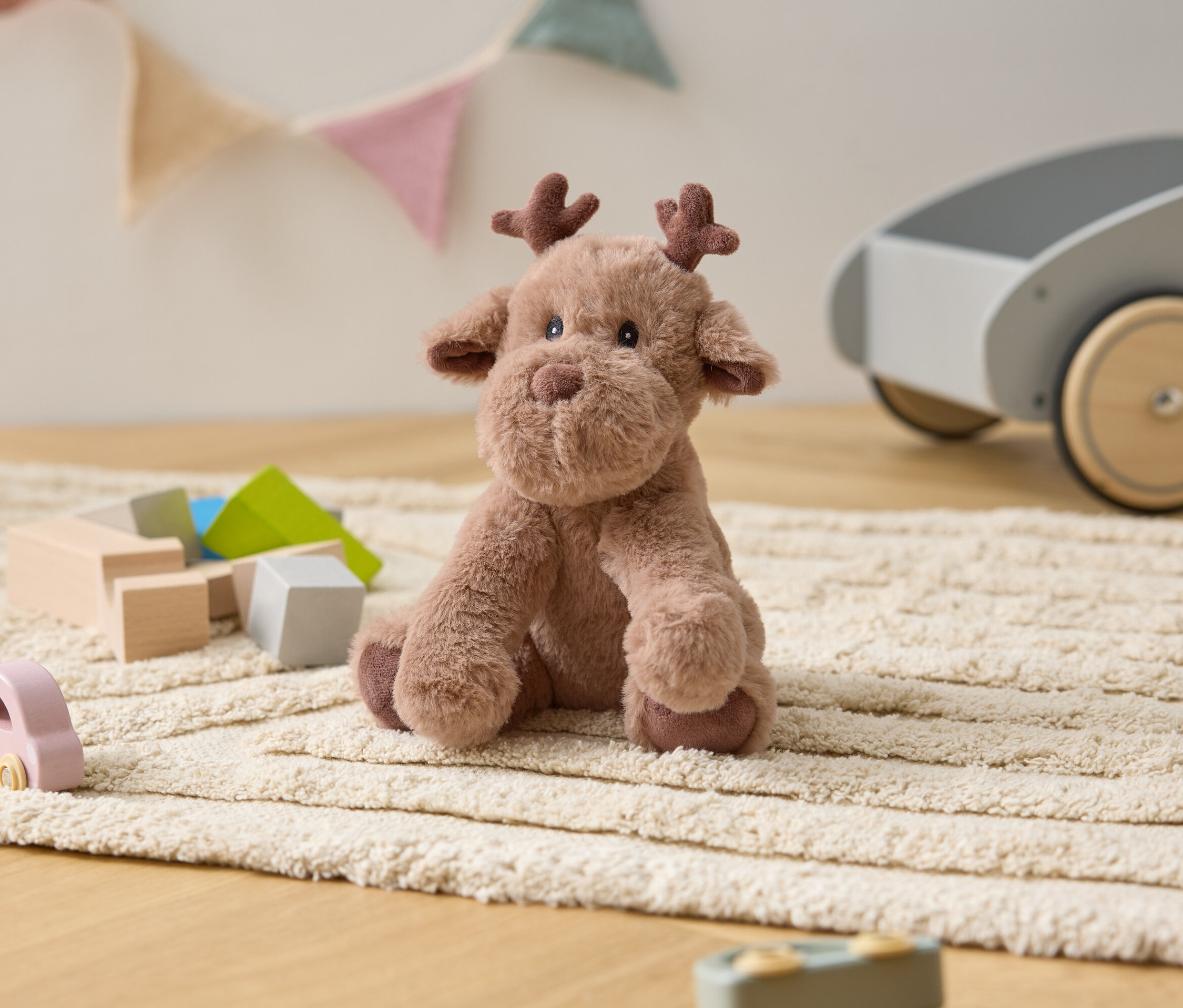 Ein Plüsch-Elch sitzt auf einem cremefarbenen Teppich zwischen Holzspielzeug und einem grauen Spielzeugwagen.