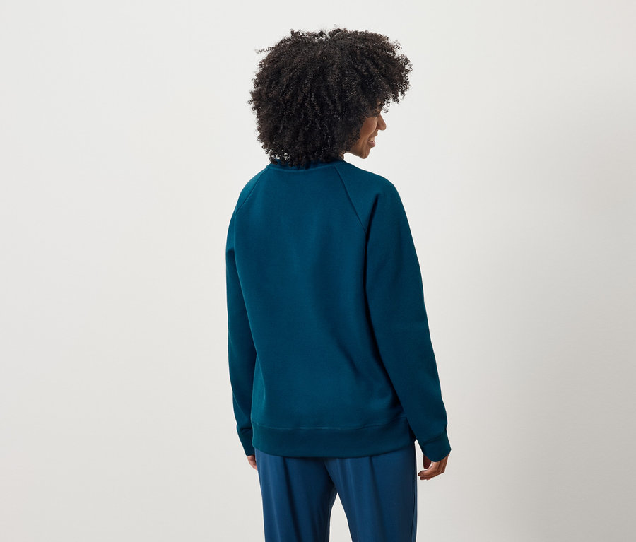 Rückansicht einer Frau mit lockigem Haar, die einen blauen Pullover und eine Hose trägt.