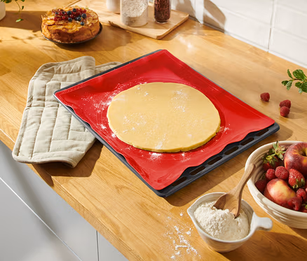 Auf einer Küchentheke liegen eine rote Dauerbackmatte mit Teig, ein Topflappen, eine Schüssel mit Obst und ein Kuchen.
