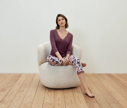 Eine Frau sitzt in einem Sessel und trägt ein violettes Shirt und eine geblümte Hose.
