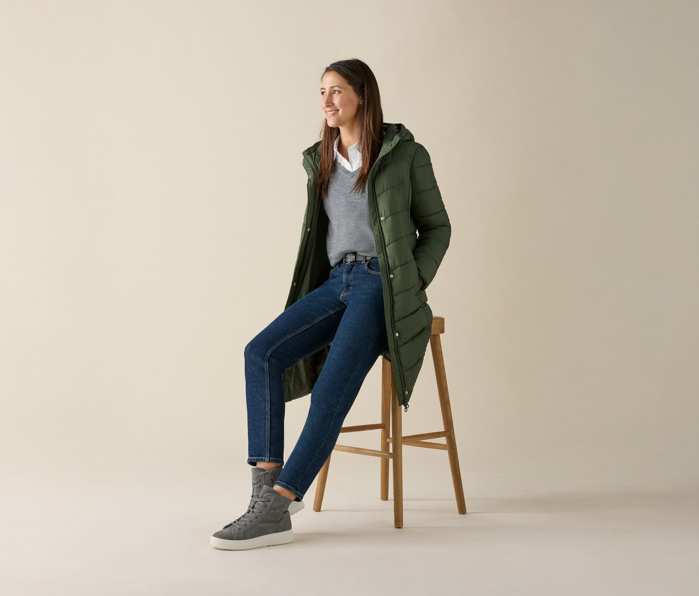 Eine Frau sitzt auf einem Hocker und trägt einen grauen Merino-Feinstrickpullover, Jeans und eine grüne Jacke.