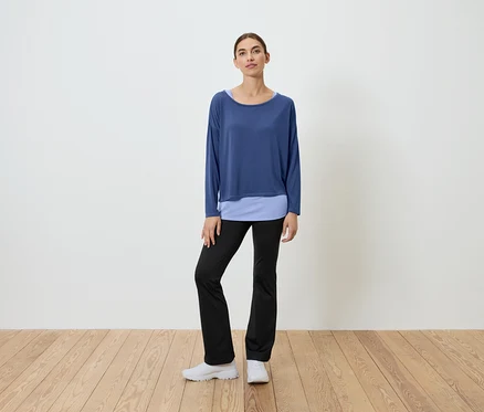 Eine Frau steht auf einem Holzboden und trägt ein blaues Oberteil und eine schwarze Hose.