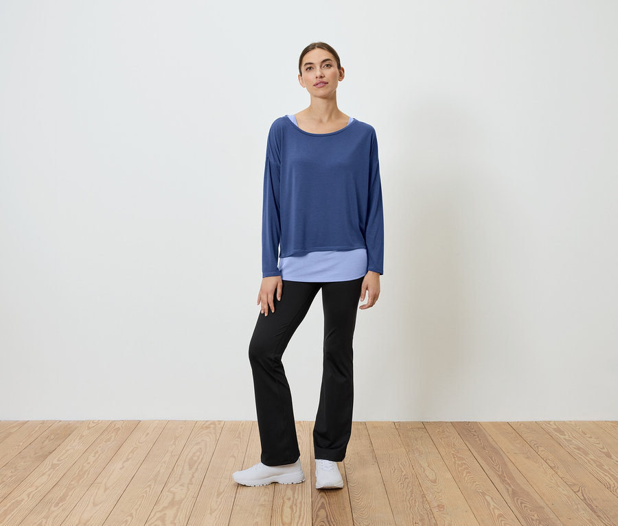 Eine Frau steht auf einem Holzboden und trägt ein blaues Oberteil und eine schwarze Hose.