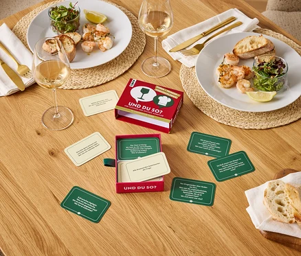 Auf einem Holztisch stehen zwei Teller mit Garnelen, Salat und Brot, zwei Gläser Wein und ein Und du so? Spiel.
