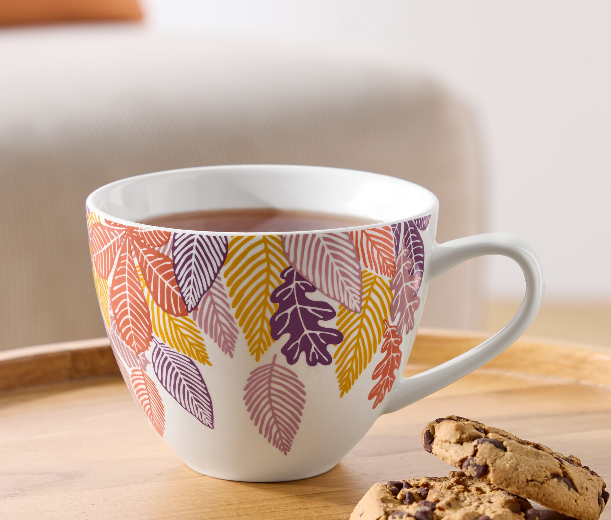 Eine Tasse Tee mit herbstlichem Blättermotiv und zwei Schokoladenkekse auf einem Holztablett.