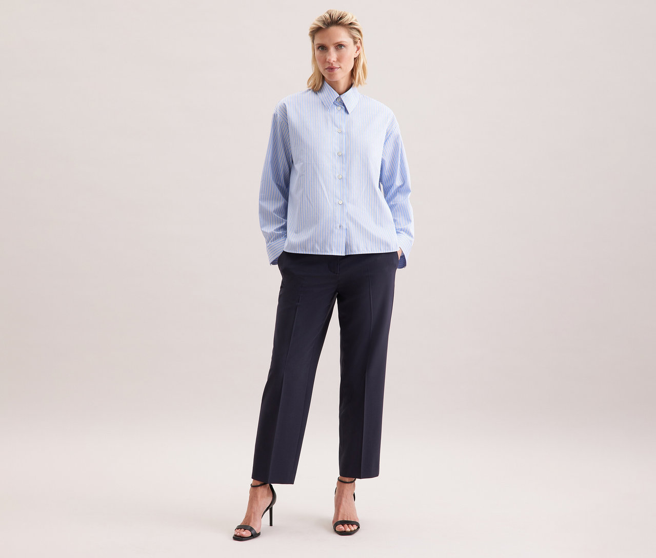 Frau trägt eine Seidensticker Oversize-Hemdbluse mit blauen Streifen, dunkle Hose und schwarze Sandalen mit Absatz.