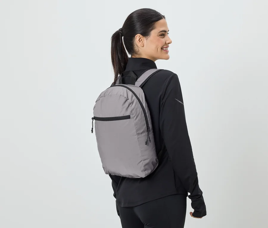 Frau mit Pferdeschwanz trägt grauen Rucksack und schwarze Sportbekleidung, Blick von der Seite.