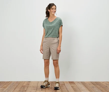 Eine Frau steht und trägt ein grünes T-Shirt, Shorts und Wanderschuhe.