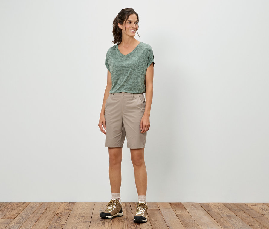 Eine Frau steht und trägt ein grünes T-Shirt, Shorts und Wanderschuhe.