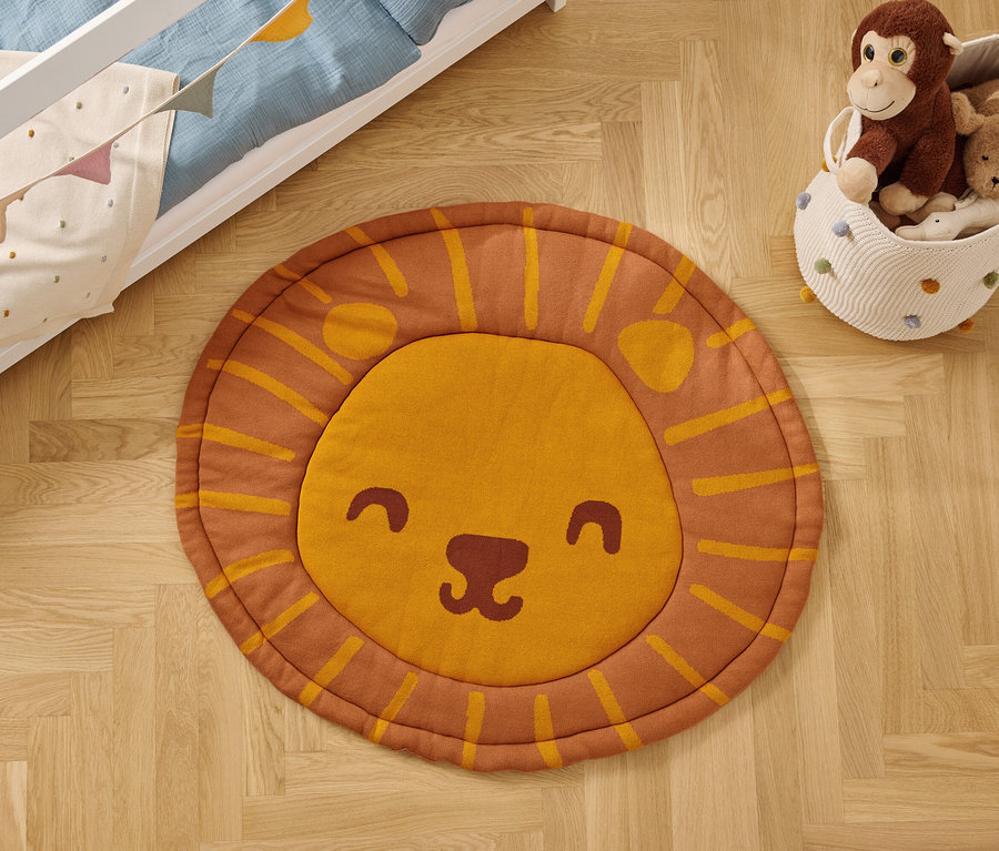 Auf einem Holzboden liegt ein runder Teppich mit einem Löwengesicht, neben einem Spielzeugkorb und einem Kinderbett.