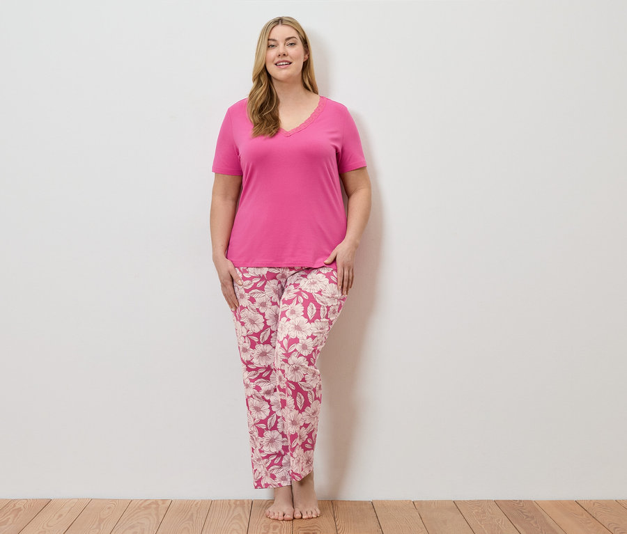 Eine Frau steht barfuß vor einer weißen Wand und trägt einen pinken Pyjama mit Blumenmuster.