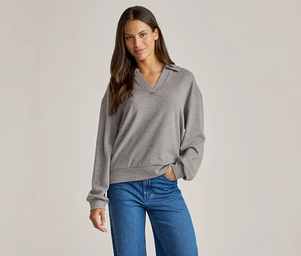 Frau mit braunen Haaren trägt einen grauen Pullover mit Kragen und blaue Jeans, eine Hand in der Hosentasche.