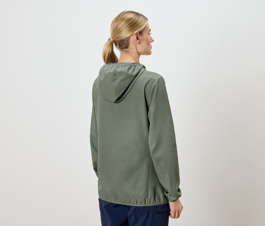 Rückansicht einer blonden Frau mit Pferdeschwanz, die eine olivgrüne Kapuzenjacke und marineblaue Hosen vor einem hellen Hintergrund trägt.