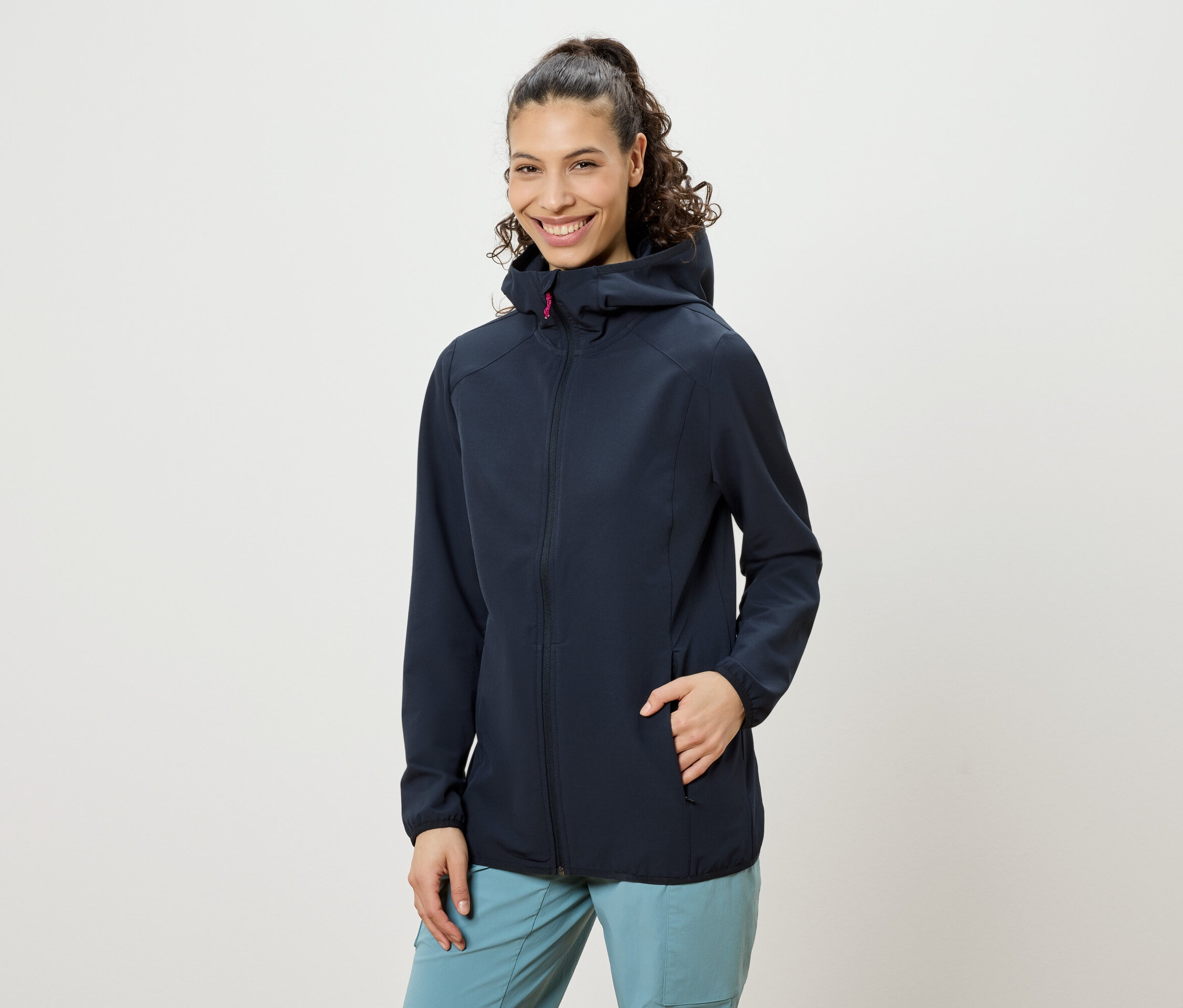 Frau mit hochgesteckten lockigen Haaren, die eine marineblaue Jacke mit Kapuze und blaue Hosen trägt und lächelt, wobei eine Hand in der Jackentasche steckt.