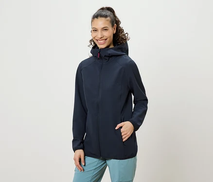 Frau mit hochgesteckten lockigen Haaren, die eine marineblaue Jacke mit Kapuze und blaue Hosen trägt und lächelt, wobei eine Hand in der Jackentasche steckt.