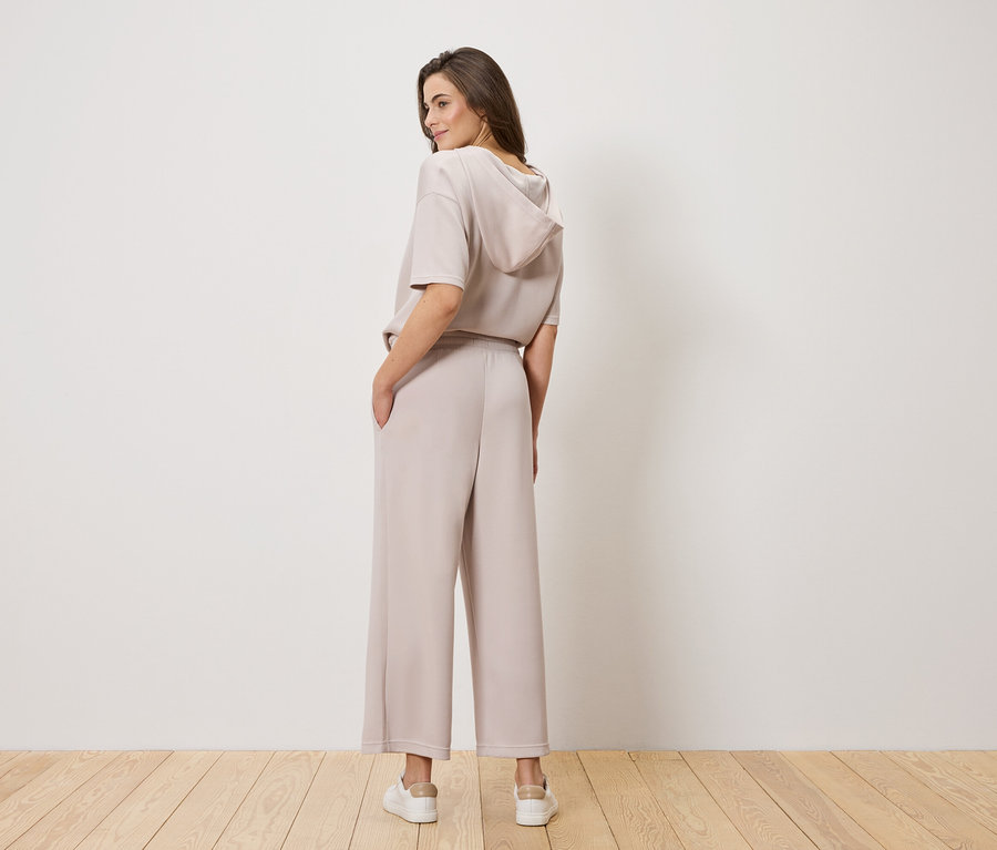 Eine Frau mit braunen Haaren, von hinten fotografiert, trägt ein hellbraunes Kapuzenoberteil und eine weite Hose.