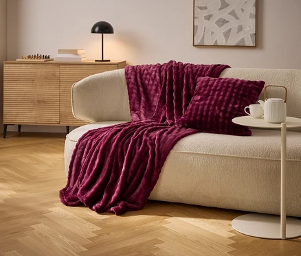 Ein burgunderrotes Plaid und Kissen liegen auf einem cremefarbenen Sofa. Ein Beistelltisch mit Teekanne und Tasse steht daneben.