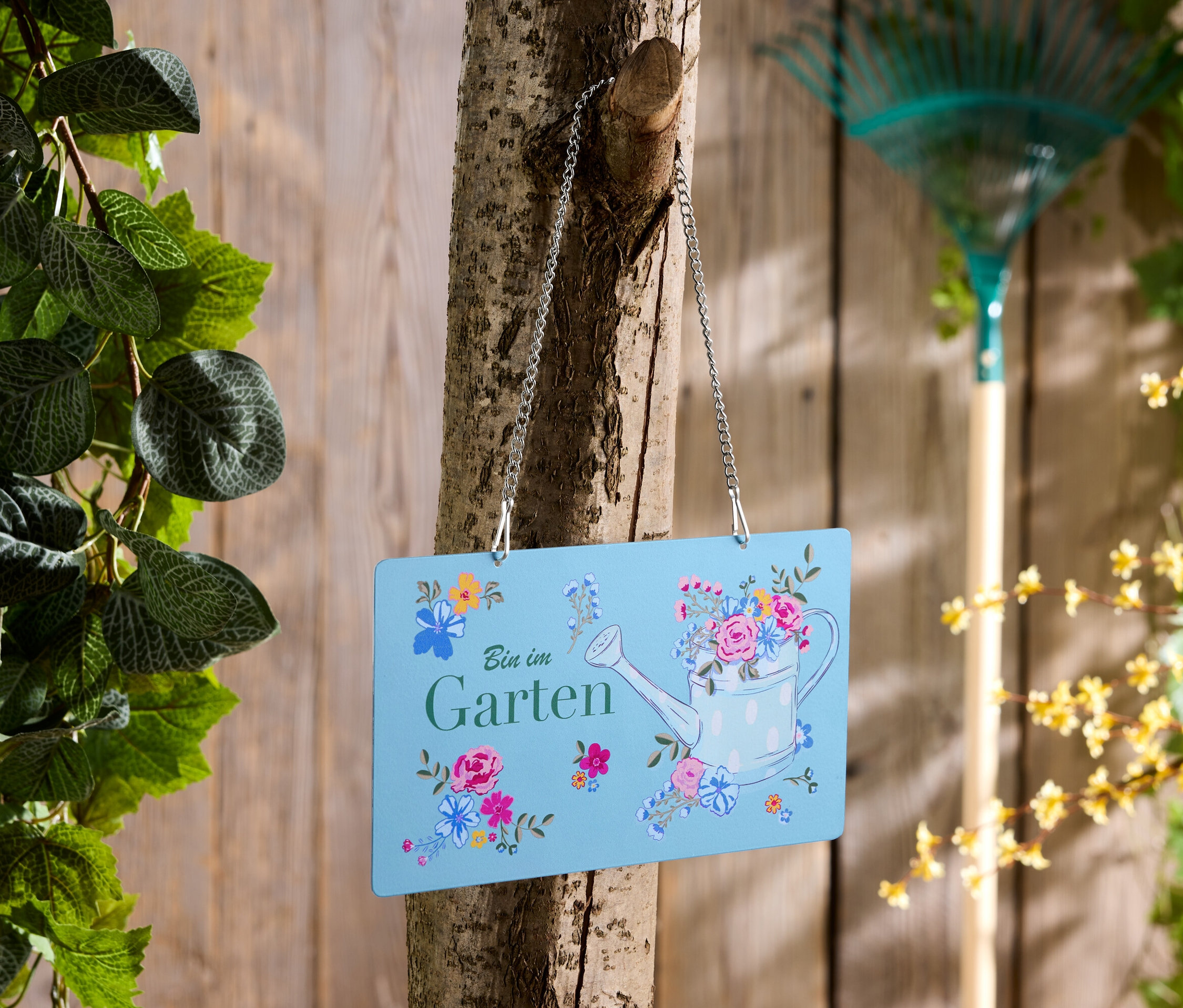 Ein blaues Schild mit der Aufschrift "Bin im Garten" hängt an einem Baum, im Hintergrund ein Rechen.