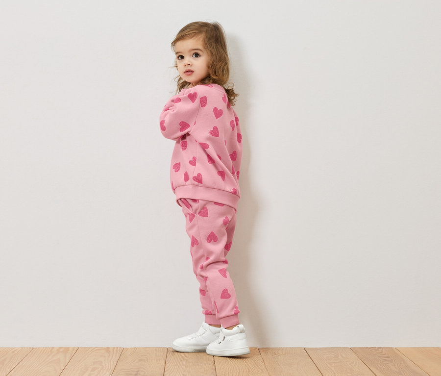 Ein kleines Mädchen steht seitlich und trägt ein rosa Sweatshirt und eine Hose mit Herzmuster, dazu weiße Schuhe.