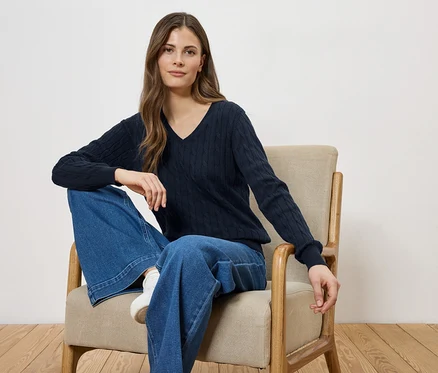 Frau sitzt in einem Sessel, trägt einen marineblauen V-Ausschnitt-Pullover und weite Jeans.