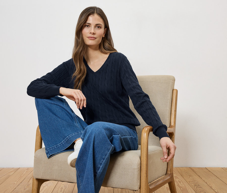 Frau sitzt in einem Sessel, trägt einen marineblauen V-Ausschnitt-Pullover und weite Jeans.