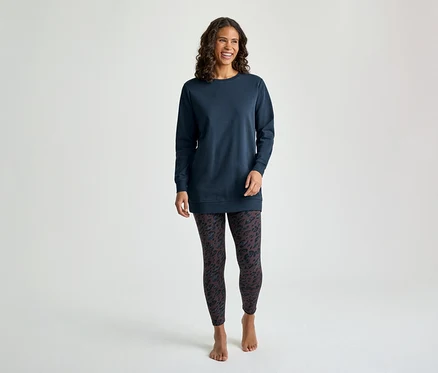 Eine lächelnde Frau mit lockigem braunem Haar steht frontal und trägt ein langärmeliges, marineblaues Tunika-Oberteil und dunkle gemusterte Leggings. Sie ist barfuß.