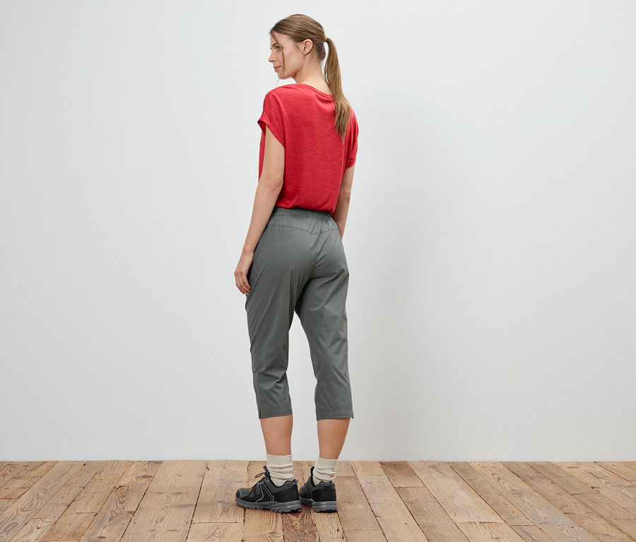 Eine Frau mit einem roten T-Shirt, grauen Hosen und Turnschuhen steht vor einer weißen Wand.