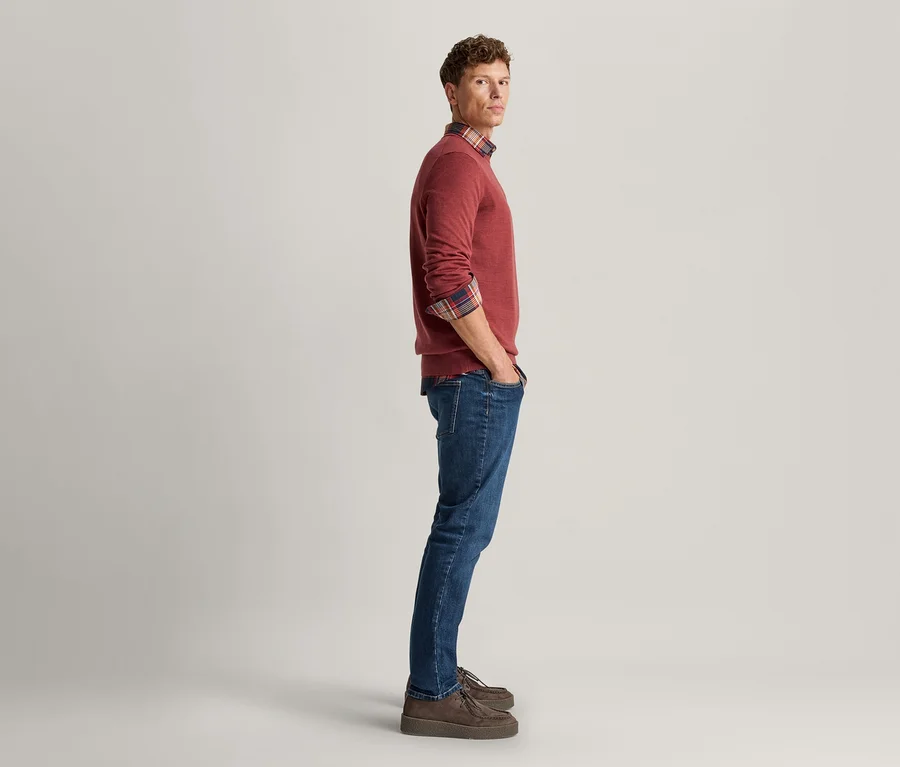 Mann steht im Profil, blickt in die Kamera, trägt einen roten Pullover über einem karierten Hemd, blaue Jeans und braune Schuhe, Hände in den Hosentaschen.