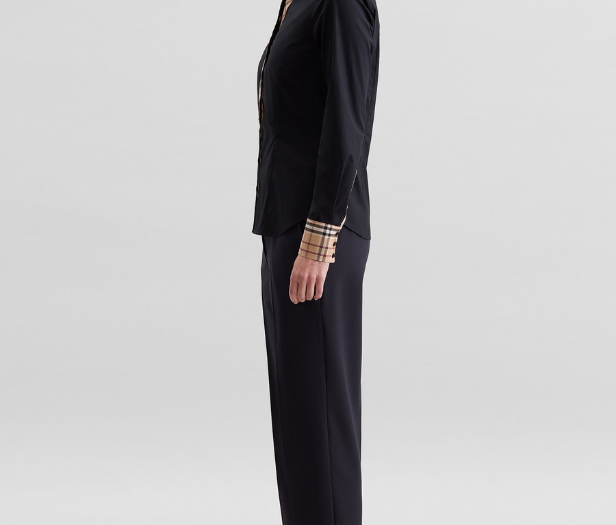 Frau im Profil, die eine schwarze Seidensticker Popeline-Bluse mit Karodetail an Kragen und Manschetten, schwarze Hose und schwarze Sandaletten trägt.